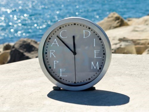 Clock with words Carpe Diem on face, sitting in front of body of water.